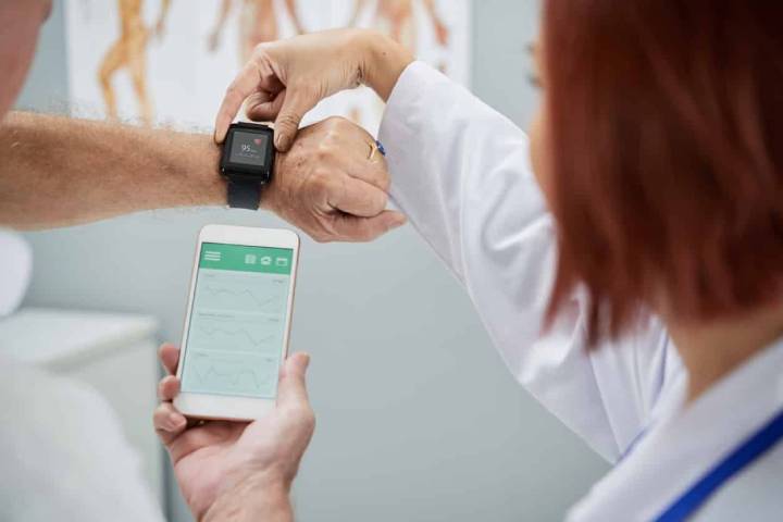 A female doctor checks the smartwatch on the patient hand and compares it to the app on a phone